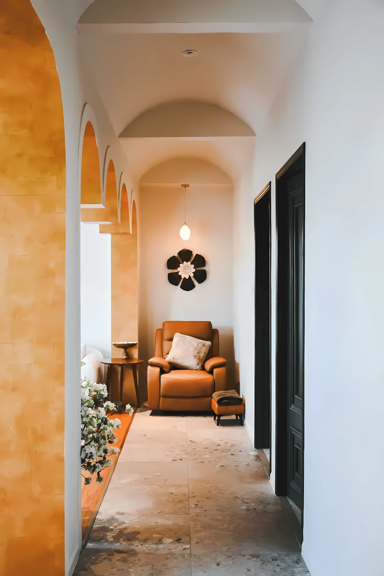 An interior view of a warm, arched hallway with ochre-colored archways on the left. The hallway leads to a cozy nook at the end, furnished with a brown leather armchair, a footstool, and a large decorative flower on the wall.