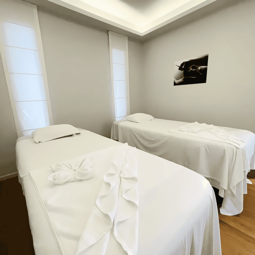 clean-couples-massage-room-white-linens Interior of a serene, minimalist spa room designed for couples, featuring two massage beds neatly made with crisp white sheets and decoratively folded towels. The room has light wood floors and modern, soft ceiling lighting.