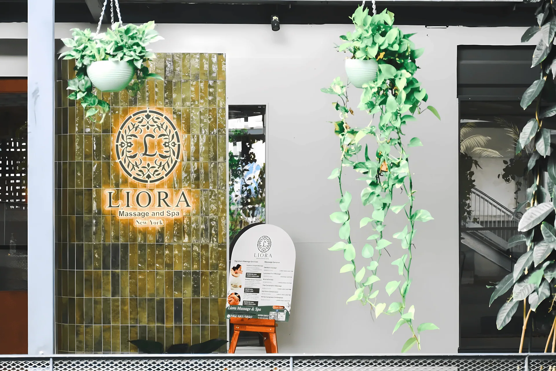 The storefront entrance of Liora Massage and Spa in New York, featuring a glowing, illuminated logo on a feature wall of glossy, earthy-green vertical tiles. A menu of services is displayed on an easel, and the entrance is framed by lush hanging plants.