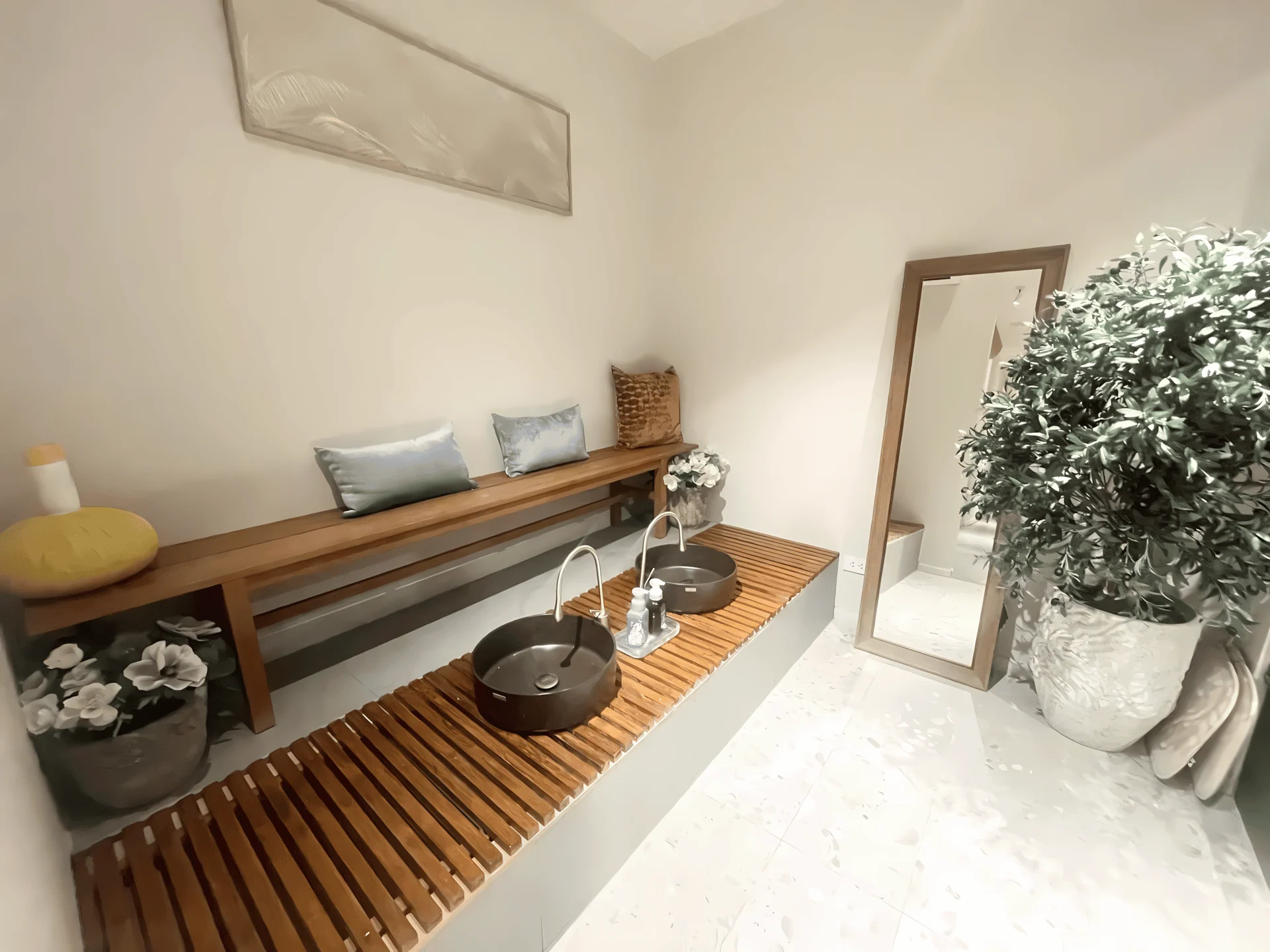 Interior of a tranquil spa foot washing station featuring two dark, modern basins on a slatted wood platform, a long bench with pillows, and decor including a large plant, mirror, and a Thai herbal compress ball.
