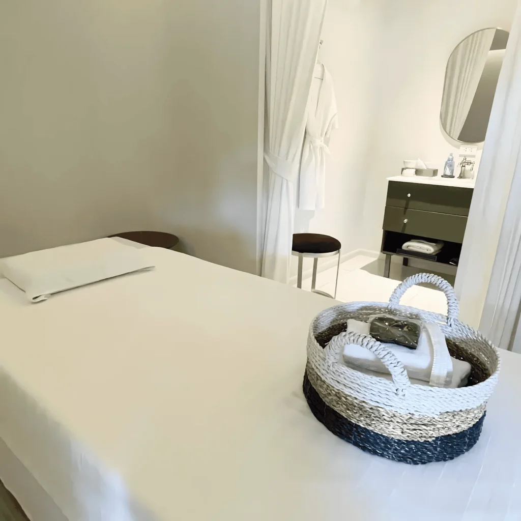 modern-spa-treatment-room-with-amenities Interior of a clean, modern spa treatment room featuring a massage table with a woven basket of fresh towels. In the background, a curtained-off private changing area includes a vanity, mirror, and a hanging bathrobe.