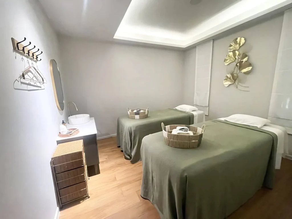 couples-massage-room-modern-spa-suite_6_11zon Interior of a modern and serene spa room for two, featuring two parallel massage beds with sage green covers, a corner vanity with a sink, light wood floors, and decorative gold leaf wall art. Baskets with fresh towels are placed on each bed.