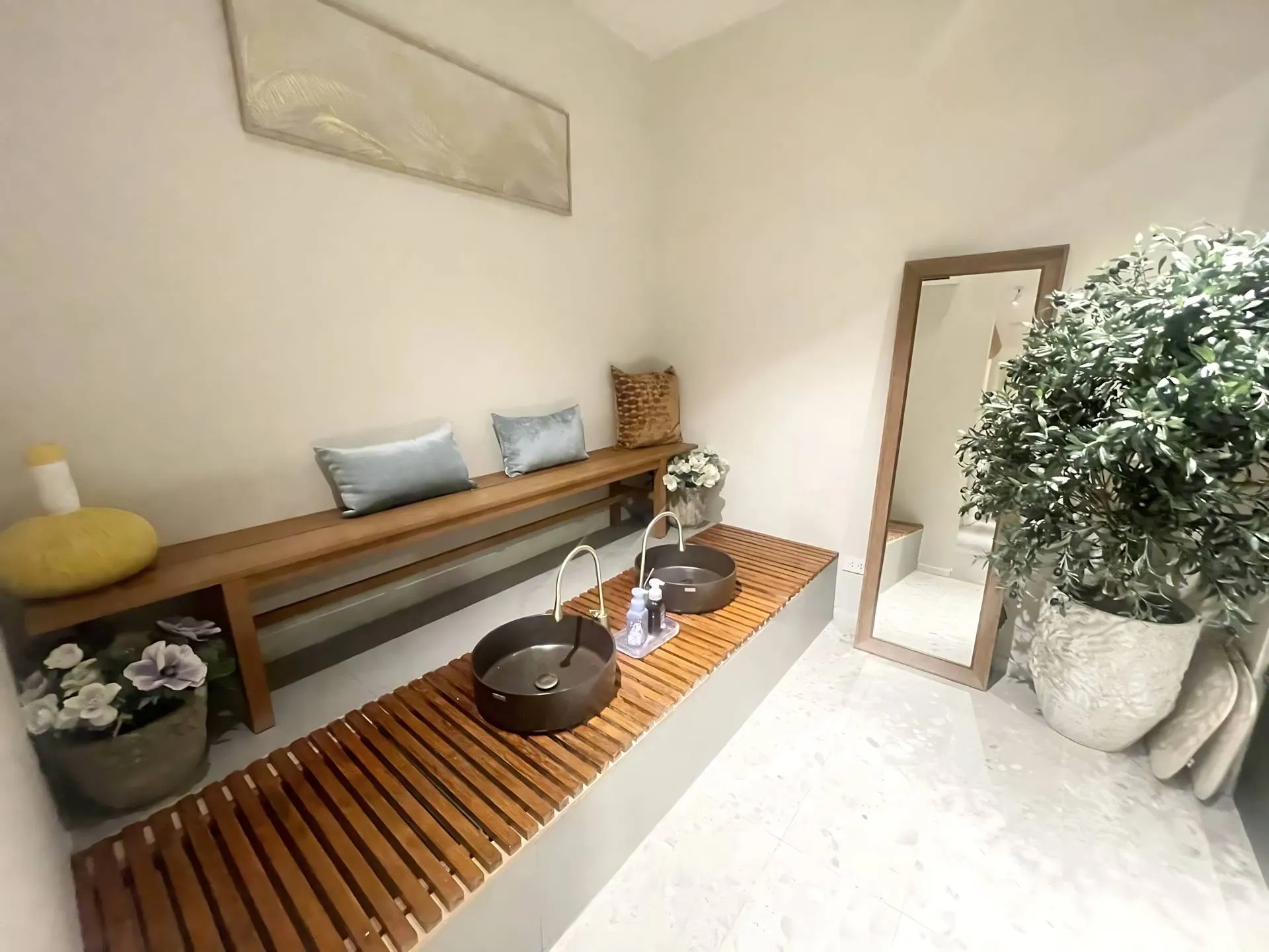 Interior of a tranquil spa foot washing station featuring two dark, modern basins on a slatted wood platform, a long bench with pillows, and decor including a large plant, mirror, and a Thai herbal compress ball.