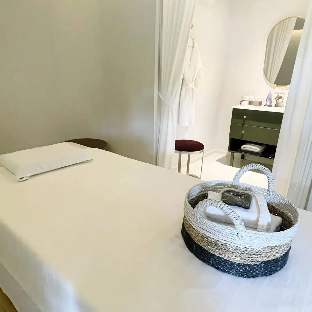 modern-spa-treatment-room-with-amenities_15_11zon Interior of a clean, modern spa treatment room featuring a massage table with a woven basket of fresh towels. In the background, a curtained-off private changing area includes a vanity, mirror, and a hanging bathrobe.