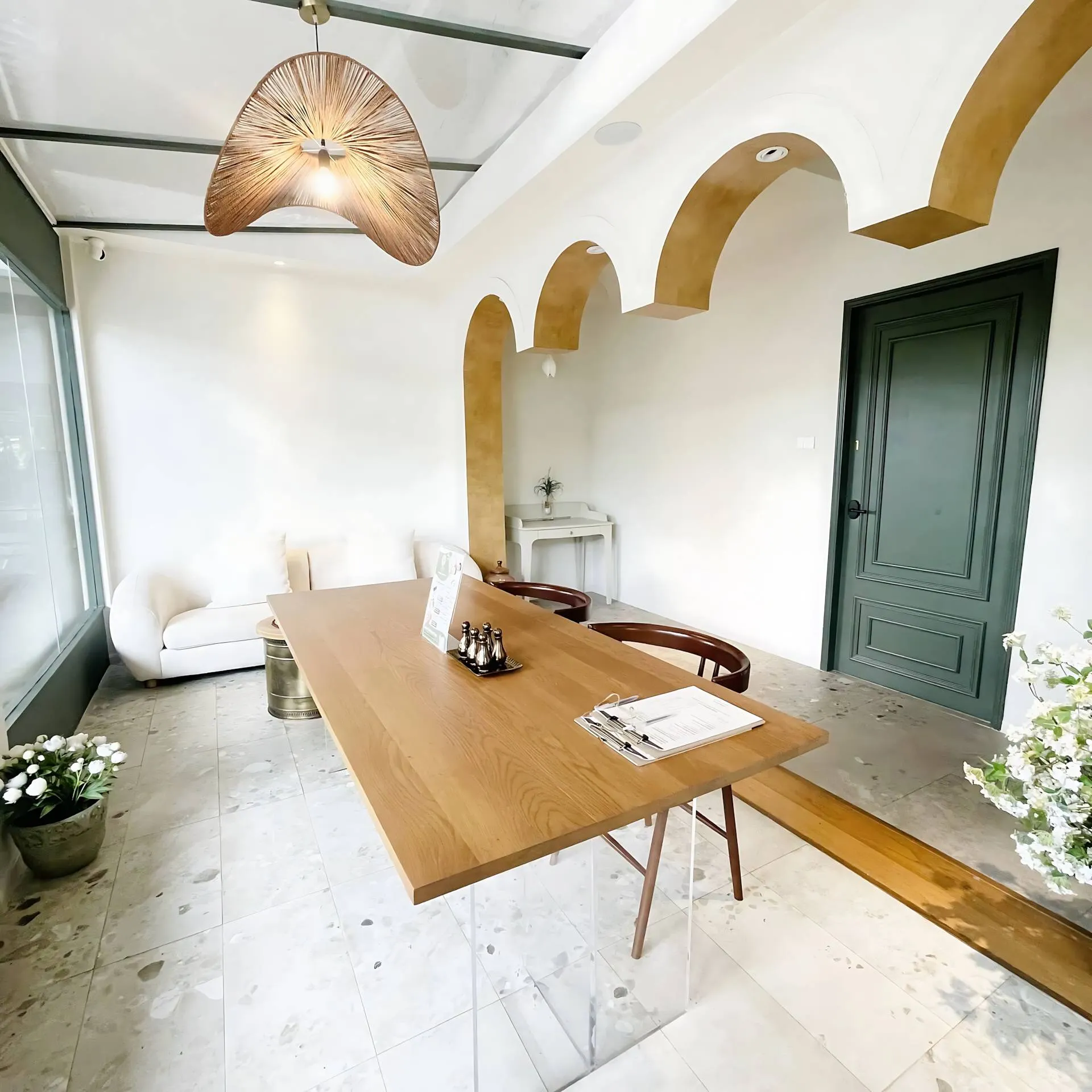 Interior of a bright, modern reception area featuring a large wood table with clear acrylic legs, a white sofa, a woven pendant light, and a distinctive wall with decorative ochre-colored arches. The floor is made of terrazzo tiles.
